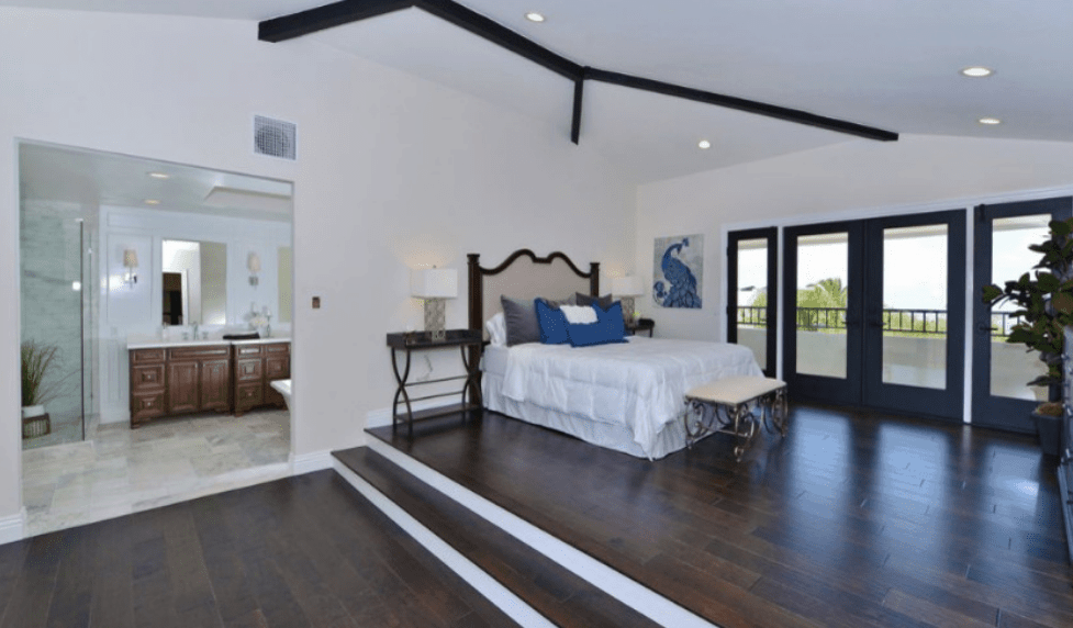 This is a stylish and spacious bedroom that highlights the dark wood with white color contrast. The dark wooden floors, exposed ceiling beams, bedside tables, and glass door boarders are all on full display. This makes the wall-mounted art, blue pillows and even the bench at the foot of the bed pop out aesthetically.