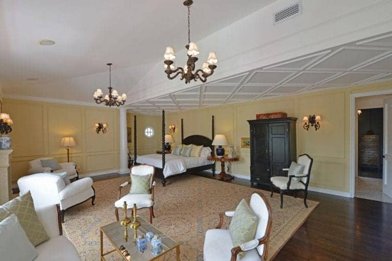 This wide Spanish bedroom includes its own living room with French chairs surrounding a glass and gold coffee table. A massive patterned rug extending from the bed to the living room area almost covers the dark wooden floors which pair perfectly with the dark wood four-poster bed. All of these are illuminated by two elegant chandeliers with matching wall-mounted lamps.