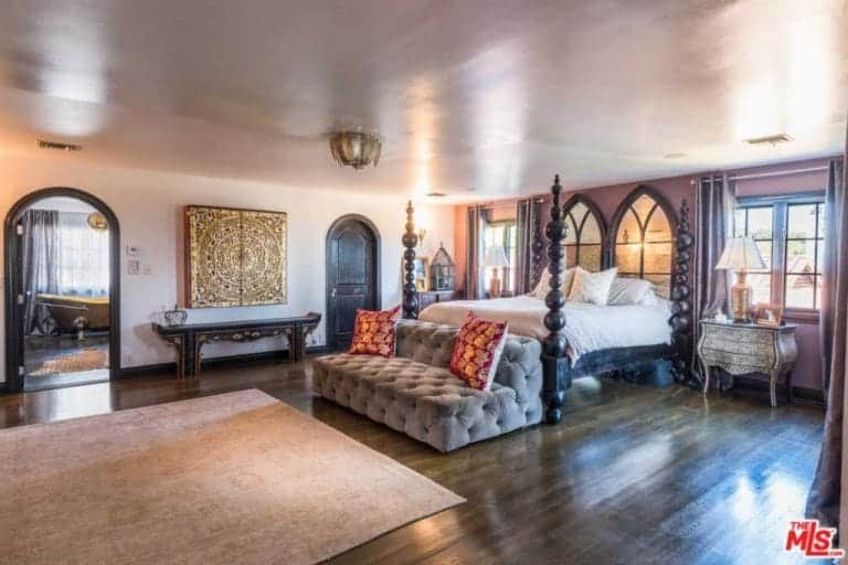 Immense Spanish primary bedroom features a huge wooden four-poster bed with elegant bead patterns at the posts. Instead of a headboard, it has a pair of Gothic-shaped windows that offer a great view of the sunset. The dark wooden floor is a great partner for the distressed side tables and brass details that are peppered across the room.