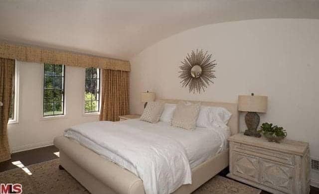The simple primary bedroom has an igloo arched ceiling that is predominantly white. This is accented with beige from the side tables, lamps and bed frame giving the room a warm neutral feel. The stand-out detail of the room is the wall-mounted gold art at the head of the board that resembles the sun or crucifix.