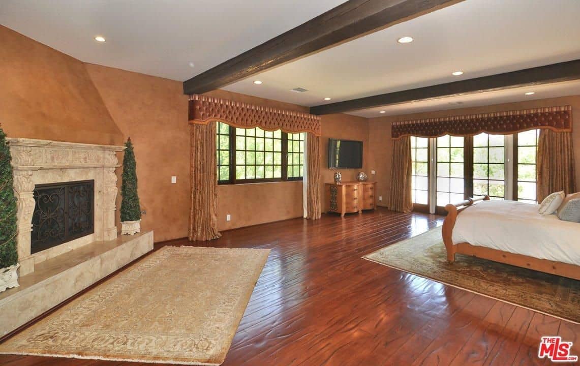 A spacious and minimalistic Spanish bedroom with a pair of exposed wooden ceiling beams. The brown walls give emphasis on the Victorian fireplace with marble stonework and iron fittings giving the room a warm and cozy feel. The pair of patterned rugs give texture to the hardwood floors and mirror the stylish curtain fittings above the windows.