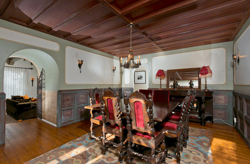 This traditional Spanish dining room has a dark wooden coffered ceiling with a small hanging iron chandelier. The dark wooden table with matching wooden chairs is well-rooted to tradition. On the other hand, this tone is balanced by a colorful patterned rug that pairs with the light hues and pattern of the wall finishing. 