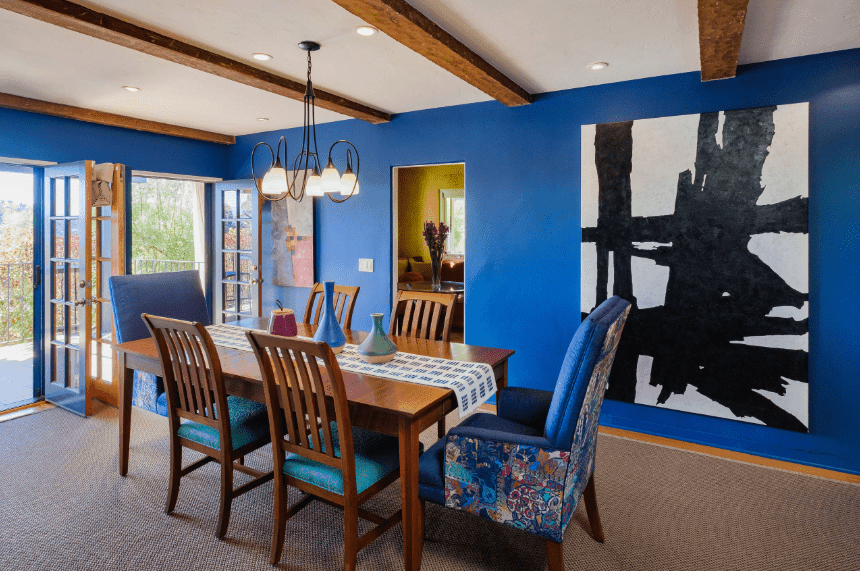 This is a predominantly blue Spanish dining room with its blue walls, French doors, and cushions on the chairs surrounding the wooden table. This is then contrasted with wooden hues from the exposed ceiling beams, table, and slat-backs of the chairs. The stark contrast is mediated by the simple chandelier, wall-mounted artworks, and grayish carpeted floor.