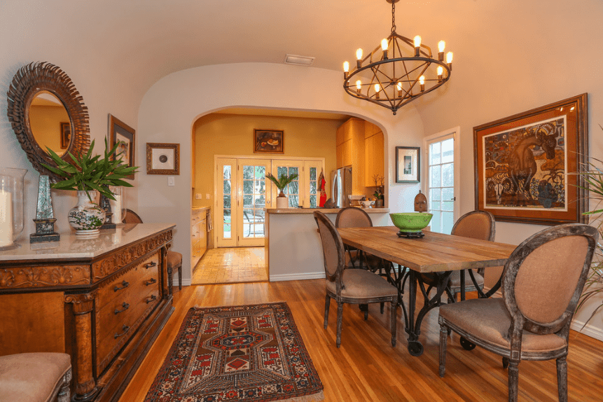 The redwood floor matches with the counter-top drawer with a circular mirror mounted above it. Across the room is a wall-mounted painting looking down on the wooden table surrounded by oval back chairs. All of these are brought together by the warm light of the black iron chandelier hanging from a white cove ceiling.
