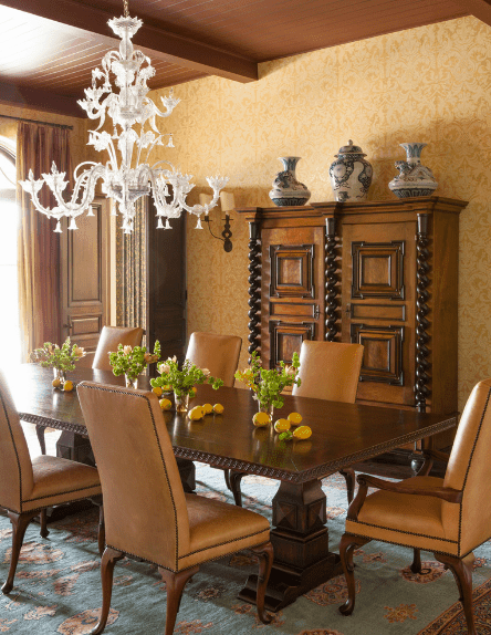 The intricate white chandelier gives contrast to this room that is dominated by the dark wooden tones of the ceiling with exposed beams, dining table, upholstered chairs, and French cabinet. The light hues of the patterned area rug reflect the vase display and patterned walls.