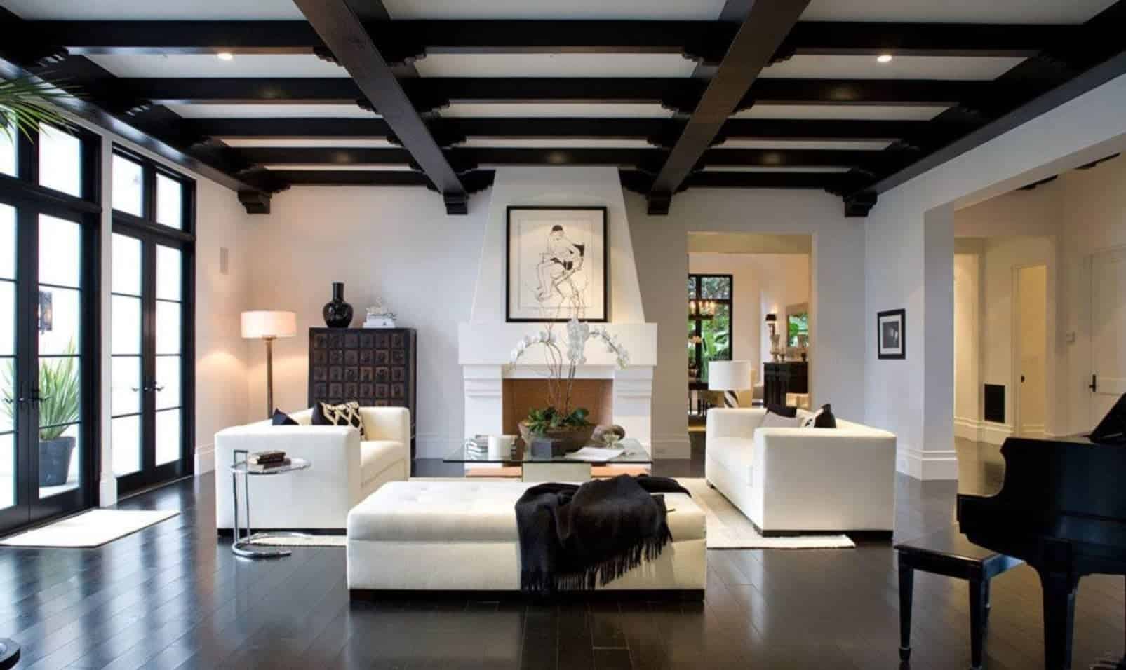 Modern white living room beautifully contrasted with a stylish black beam ceiling and hardwood flooring. It has white matching sofas and tufted bench accented with black pillows and throw blanket.