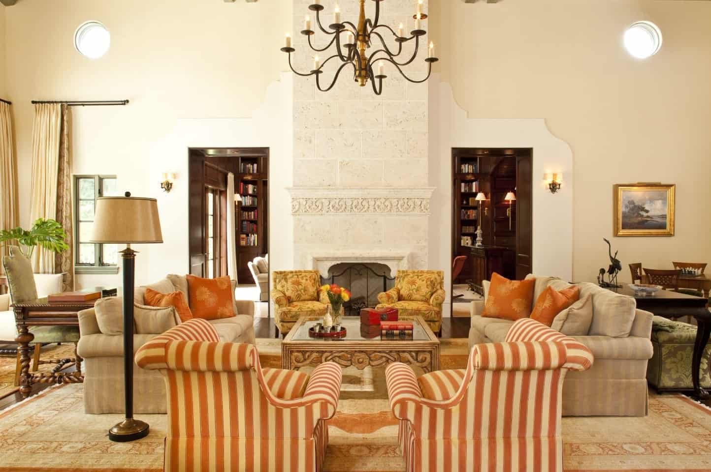 Symmetrical living room features matching floral printed and red striped chairs along with taupe facing sofas over a vintage rug. It centers around a glass top coffee table and fireplace situated in the middle of opened doors lighted by wall sconces.