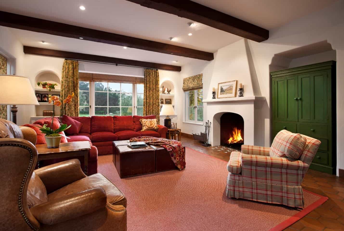 Cozy living room boasts an arched fireplace and red sectional sofa paired with an ottoman center table and mismatched chairs. It has framed glass windows overlooking the green outdoor view.