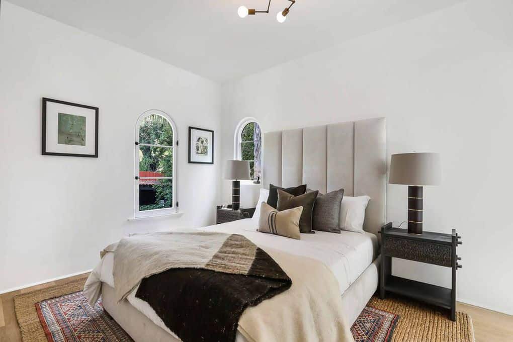 This simple white bedroom is dominated by the woven wicker rug in the middle of the floor. A pair of dark modern lamps sit atop of side tables that goes in contrast with the white walls and light gray headboard. Two arch top windows give accents to the walls along with wall-mounted photos.