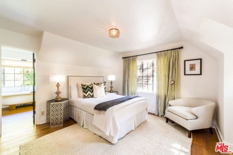 An elegantly simple white bedroom that has a white rug, white bed, white walls and white single sofa chair at the corner. There are brass details that accentuate the room elegantly like the side tables and lamps and the standing lamp by the sofa chair. These are all illuminated by a ceiling-mounted flush lighting with an intricate brass design.