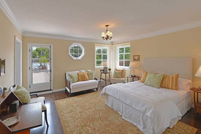 This Spanish style bedroom has a color theme of subdued yellow which is evident in the patterned rug, pillows and the color of the walls. This is brought to life by the yellow light of the bedside lamps and a small chandelier hanging in the middle of the room. Amidst all that, an octagonal window stands out above a small two-seater sofa.