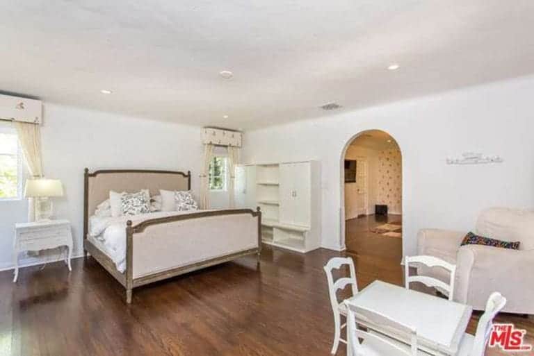 An elegant white bedroom with a beautiful arched doorway. The brown wooden floors elevate the whiteness of the walls, side table, chairs, cabinet, and even the single sofa at the corner. It gives them a semblance of floating. This allows the bed to stand out even with its muted wood and grayish brown headboard.