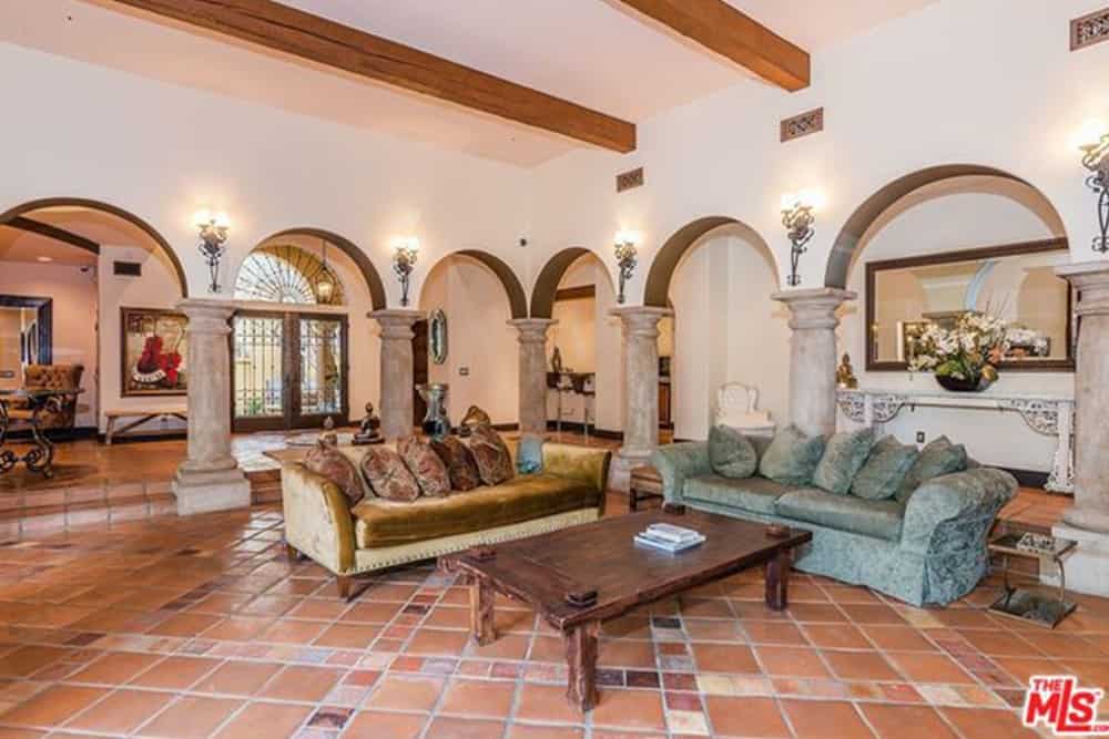 Spacious living room surrounded by open archways that are lighted by wall sconces. It has velvet brown and green adjacent sofas paired with a wooden table over terracotta flooring.