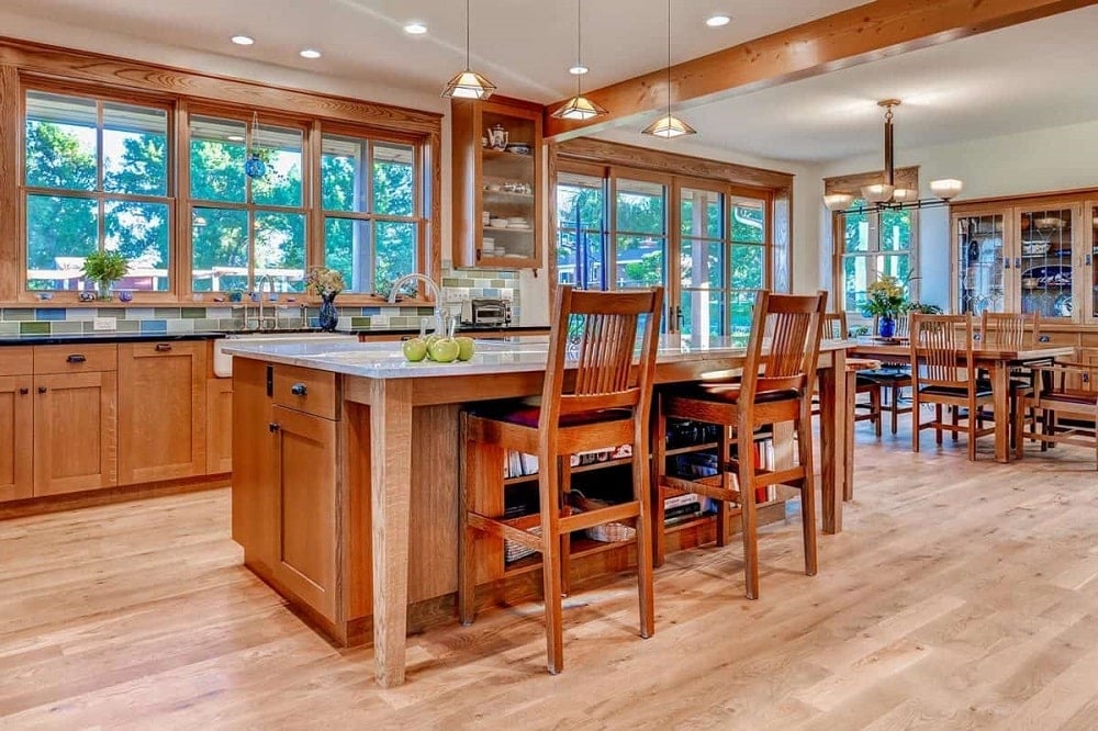 This is a close look at the kitche with consistent wooden tones on its kitchen island, stools and cabinetry complemented by the natural lighting that comes from the row of windows.