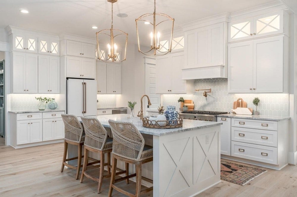 The Farmhouse-style kitchen has a large white kitchen island topped with lantern pendant lights and paired with woven wicker stools with backs.