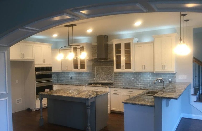 The the large arched entryway is the kitchen that has gray subway tiles to match the gray marble countertops and white cabinetry warmed by the yellow lighting and the pendant lights over the kitchen island.