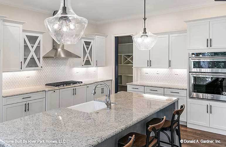 The kitchen is equipped with a double wall oven, a built-in cooktop, white cabinetry, and a breakfast island that is topped with glass pendant lights hanging from a white ceiling.