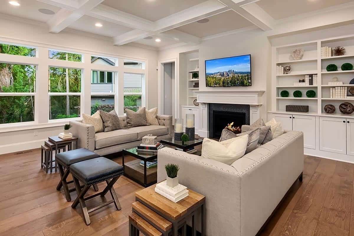 The great room is furnished with beige sofas, cushioned stools, and nesting tables. It is crowned with a coffered ceiling that's fitted with recessed lights.