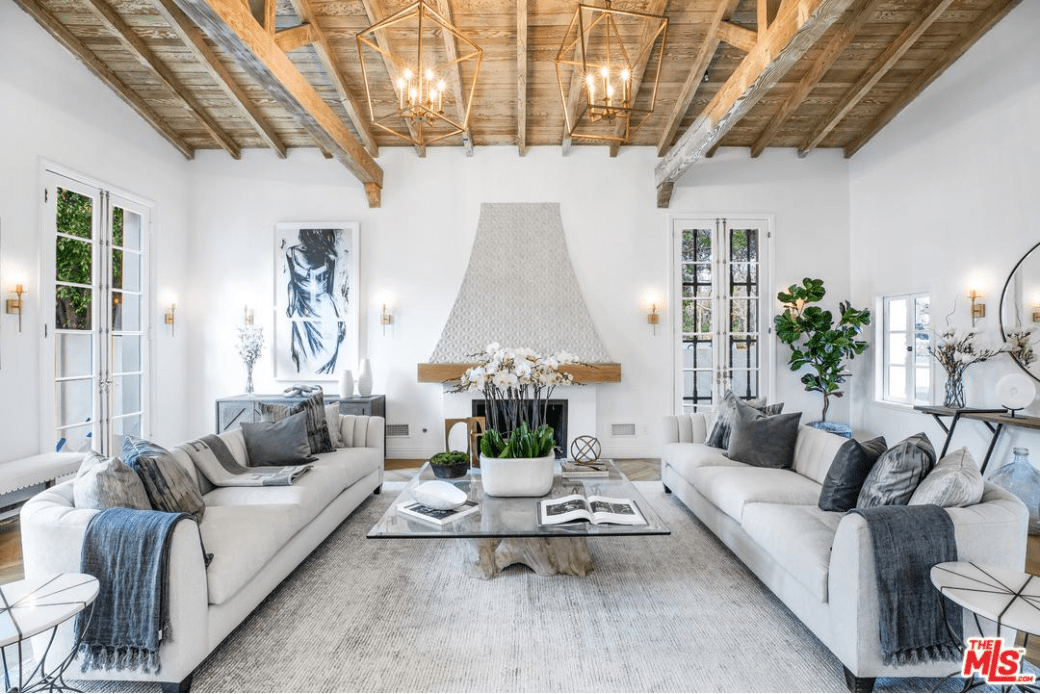 Bright living room lighted by a pair of gold industrial chandeliers and sconces surrounding the white walls. It features gray sectionals facing each other with a tree stump coffee table topped with glass.