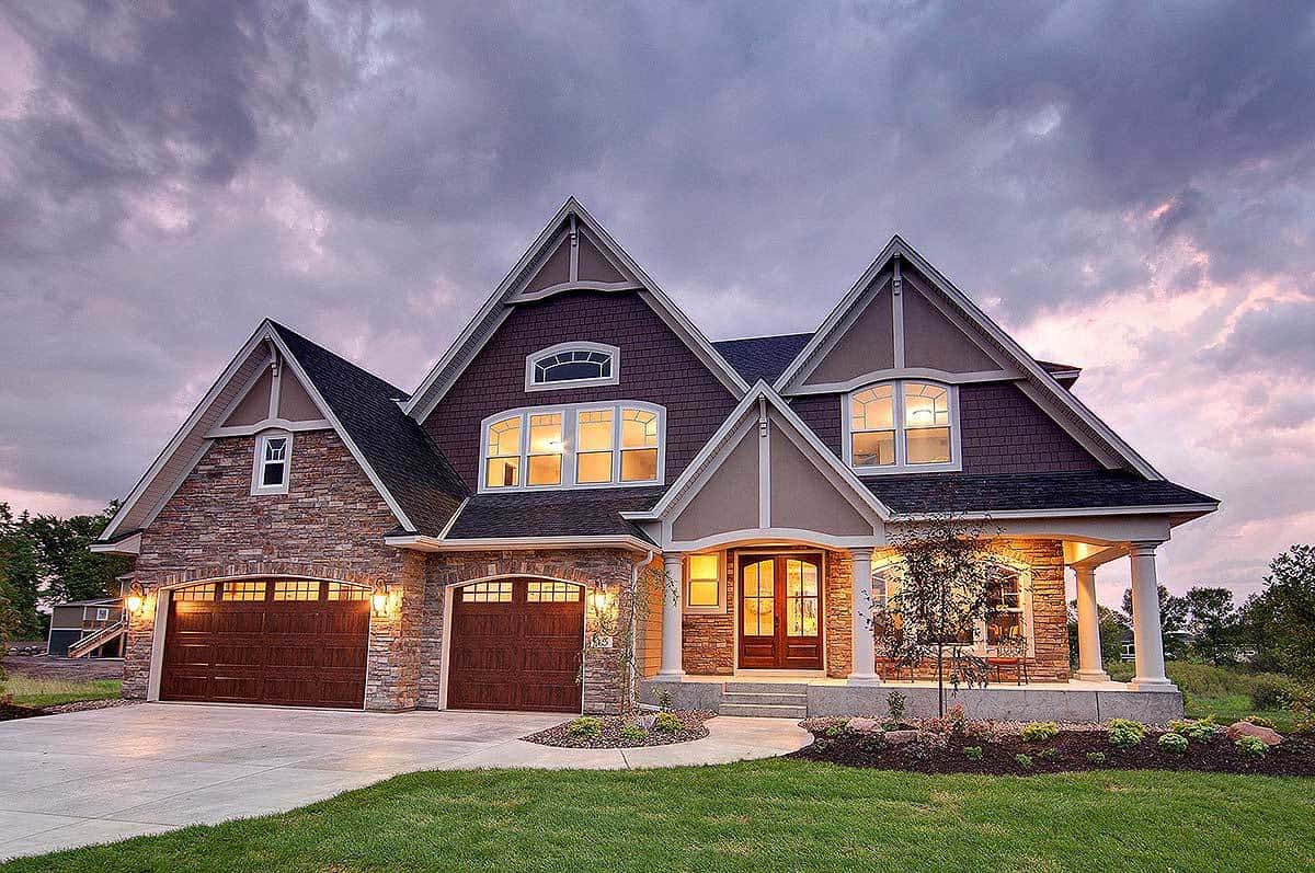 The warm yellow glow of the windows matches with the outdoor lamps on the exterior walls accented with stone. This is then complemented by the simple grass lawn with a medium-sized tree and the wide concrete driveway beside it.