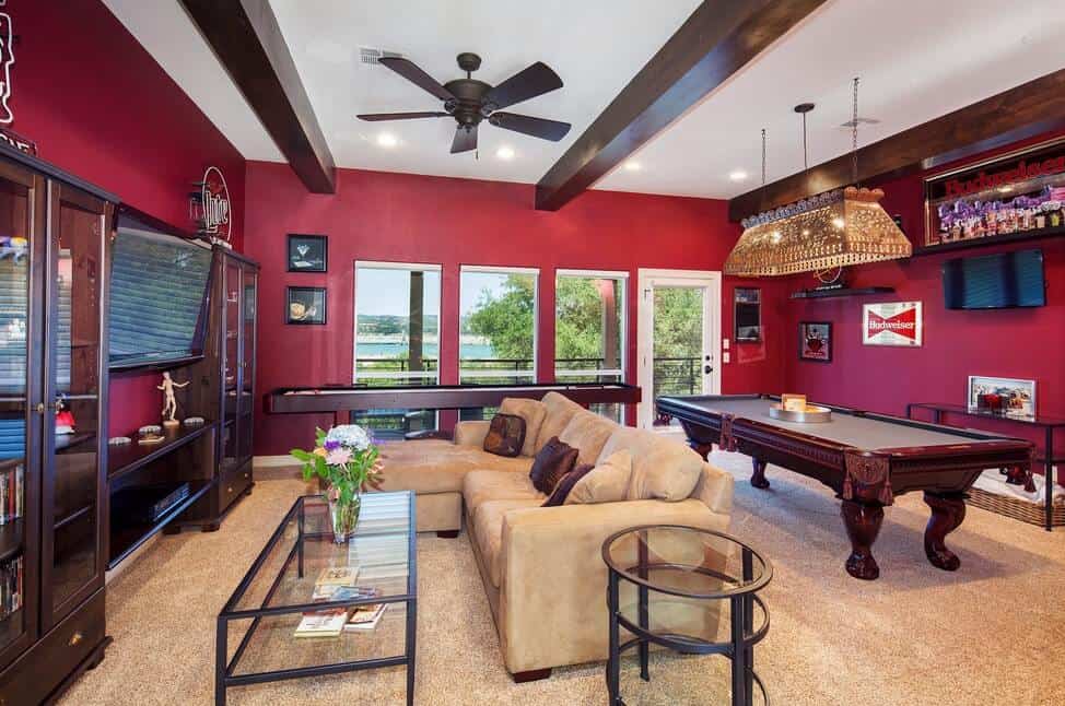Red family room showcases flat panel TV in between glass front cabinets and tan sectional complementing with the carpet flooring. It is accompanied by glass tables and a pool table at the back side lighted by an ornate linear chandelier.