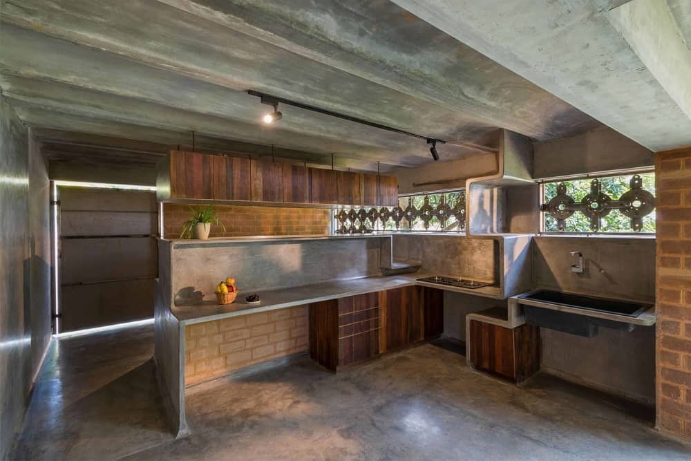 This is a full view of the kitchen with concrete structures on the walls along with wooden cabinetry. These are complemented by the concrete beams of the ceiling and the patterned panels of the windows.