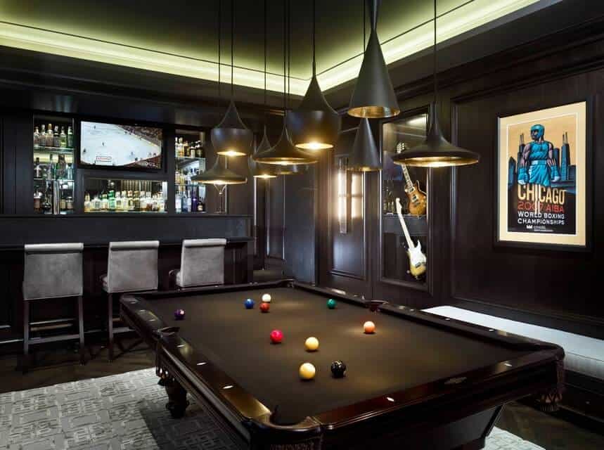 Black game room designed with a wall art and guitar displays offers a bar area and black pool table lighted by various pendants that hung from the luminous tray ceiling.