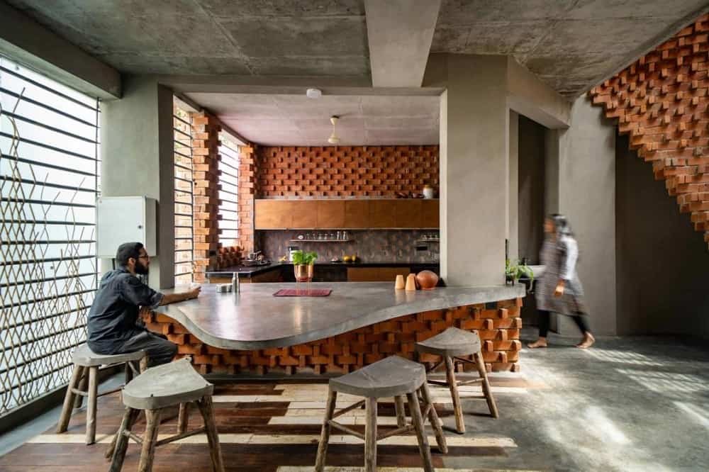 This is a full view of the kitchen that has a large concrete bar with concrete pillars paired with wooden stools for the breakfast bar. On the far side are the wooden cabinetry lining the walls adorned by the filtered lighting and red brick textured walls.