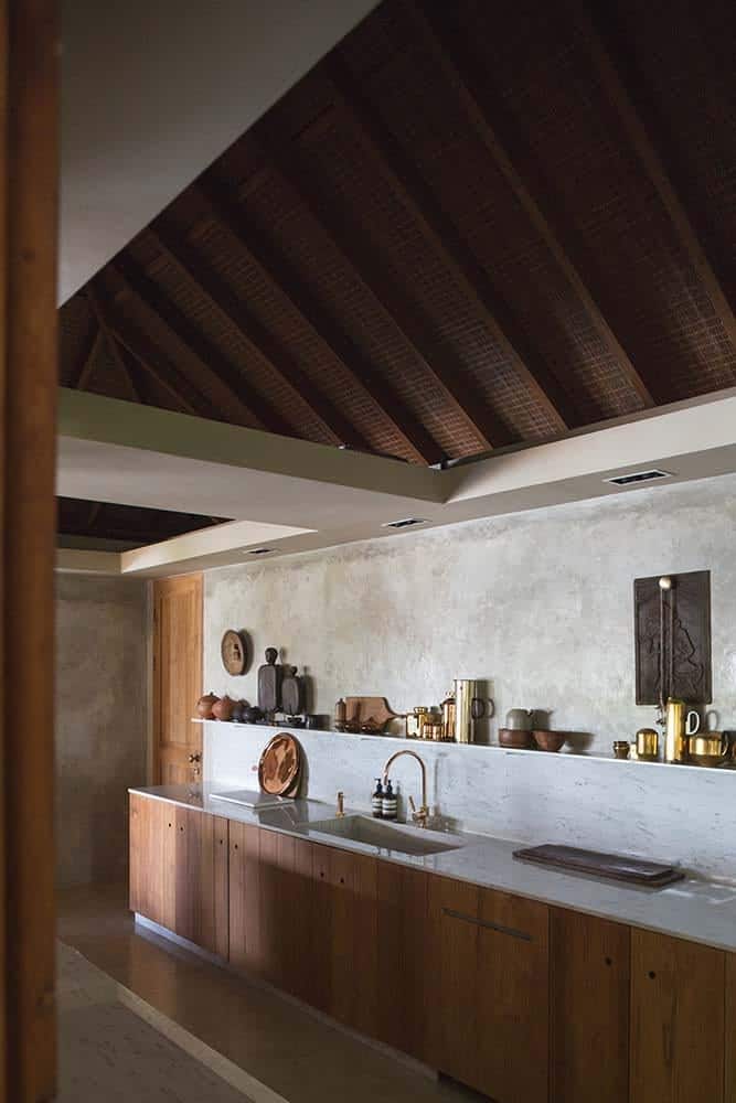 This is a close look at the kitchen that has a dark wooden beamed ceiling with an arch over the concrete walls and structures. These match well with the wooden cabinetry topped iwth bright counters and backsplash that has a built-in shelf.