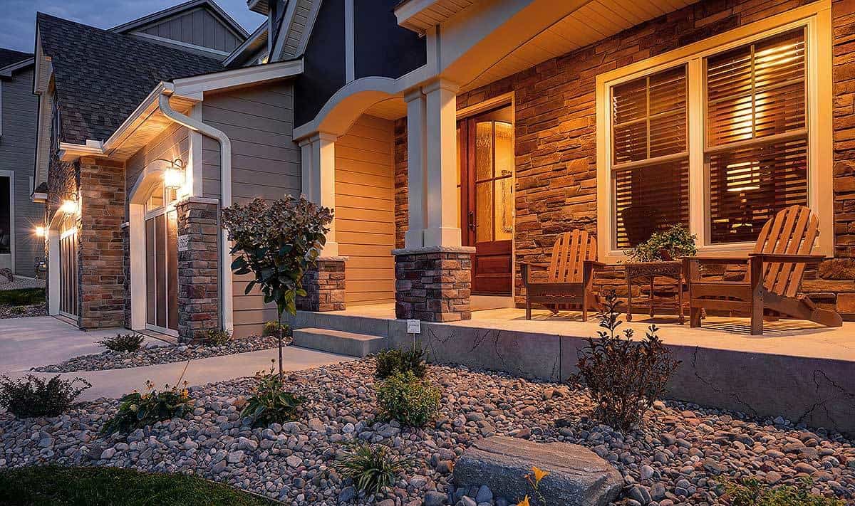 This is a closer look at the front of this lovely house with a pebbled plot next to the grass lawn. This small area has small trees and shrubs illuminated by the warm glow of the front porch.