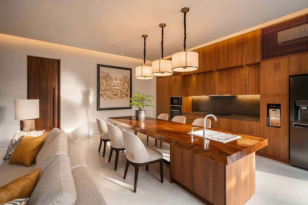 This is the eat-in kitchen with a large wooden kitchen island that has an attached table topped iwth drum pendant lights. These are then paired with wooden cabinetry on the far wall that makes the appliances stand out.