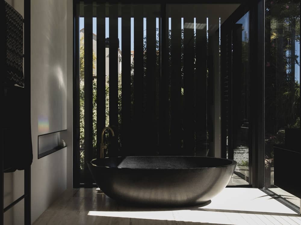 This is a black bathroom with black wooden slat panels and a matching black freestanding bathtub that is brightened by the glass window.