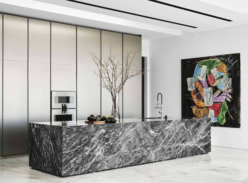 This is a full view of the modern kitchen that has a large black and white marble kitchen island that stands out against the white marble flooring and modern white cabinetry on the other side that houses the modern appliances. These are then complemented by the large colorful wall artwork.