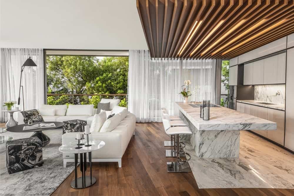 This is a close look at the kitchen on the side of the living room with a white marble kitchen island topped with a wood beamed ceiling and modern sleek white cabinetry on the far wall.