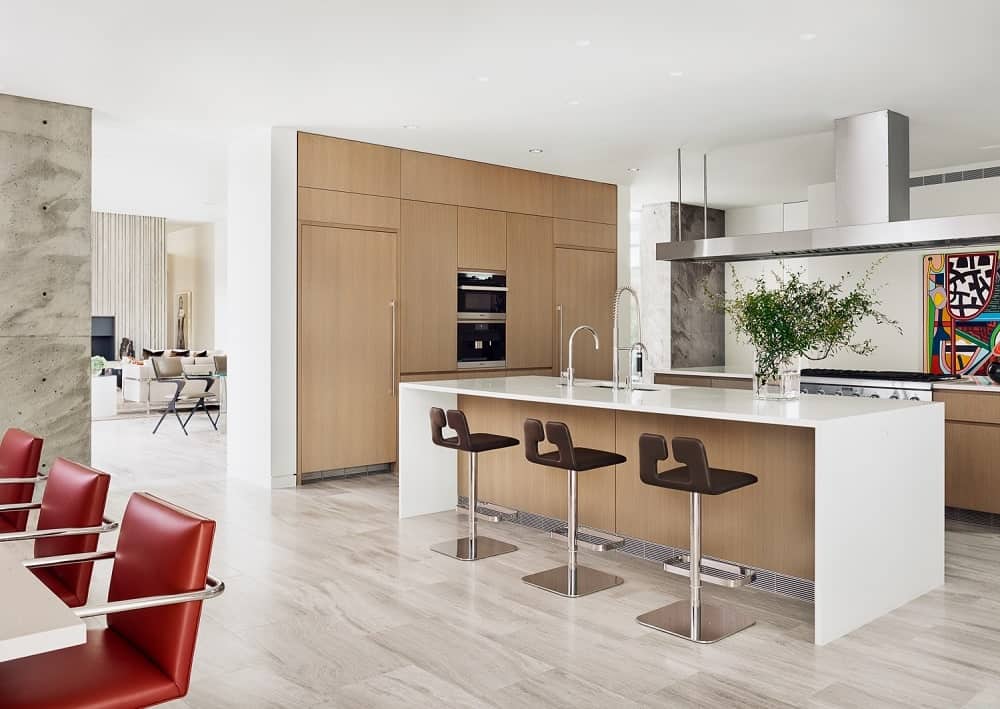 The kitchen has a white waterfall kitchen island paired with dark stools for its breakfast bar paired iwth a modern wooden cabinetry on the wall with the oven.