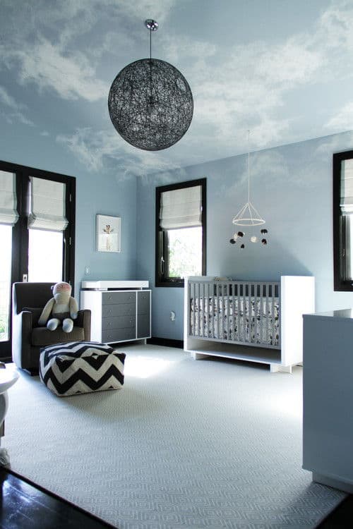 Marvelous nursey features a textured area rug and a black spherical chandelier that hung from the vaulted ceiling painted with serene sky mural. It includes a white crib and gray armchair paired with chevron ottoman.