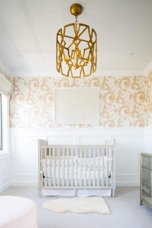 A stylish gold chandelier adds a stunning accent in this nursery clad in peach swirl wallpaper and white wainscoting. It has a natural wood crib and a white faux fur rug that lays on the gray carpet flooring.
