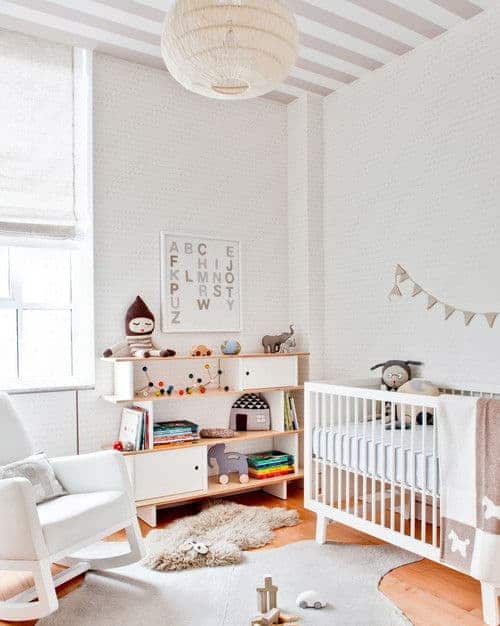 White nursery decorated with alphabet wall art and a spherical woven chandelier that hung from the striped ceiling. It has a white crib and a cushioned rocking chair over layered rugs and hardwood flooring.