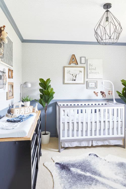 Fresh nursery boasts gallery walls and a cowhide rug that lays on the carpet flooring. It includes geometric chandelier and a white crib flanked by green potted plants.