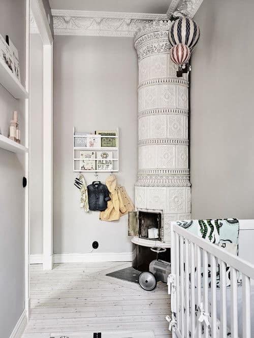 A gorgeous corner fireplace designed with intricate details is the highlight in this baby nursery with floating shelves and a white crib over light wood plank flooring.