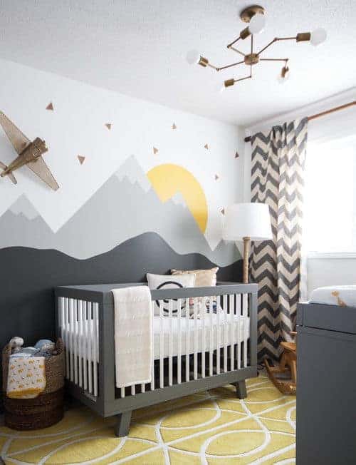 This nursery showcases chevron drapes and a gray crib with a mountain mural backdrop. It is illuminated by a brass chandelier and a traditional floor lamp.
