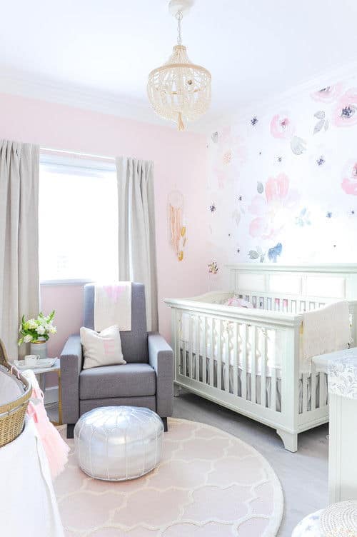 Charming nursery with a white crib and gray armchair paired with a silver ottoman over a round patterned rug. It is lighted by a boho chandelier and natural light from the picture window covered in blackout draperies.