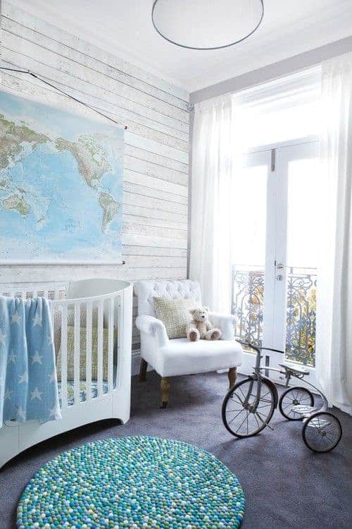 Natural light streams in through the French door that opens to the balcony framed with ornate wrought iron railings. This nursery has a gorgeous round rug and a tufted armchair next to the white crib under a map tapestry.