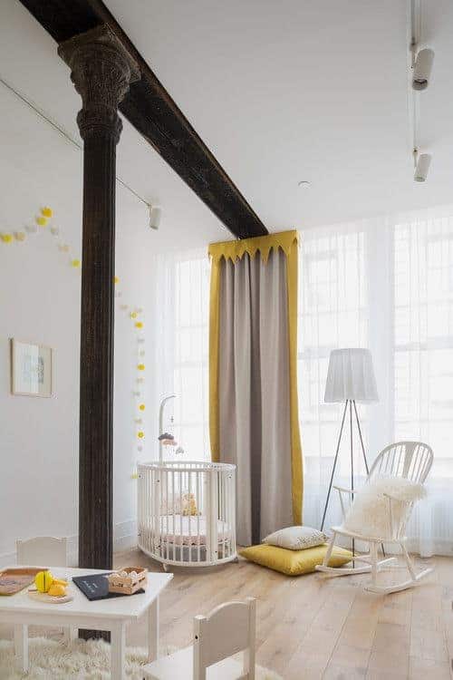 Yellow valance, pillow and pompom garland adds a pop of color in this neutral nursery lined with a vintage column. It has a white table and crib that matches the rocking chair lighted by a tripod floor lamp.