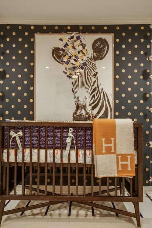 A black dotted wall mounted with a zebra portrait sets a gorgeous backdrop to the wooden crib that sits on a carpet flooring topped by a striped runner.