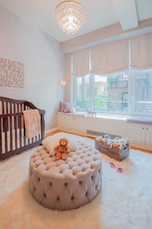 A fancy chandelier illuminates this nursery offering a wooden crib and round tufted ottoman over a white furry rug. It includes a lovely artwork and a seat nook by the glass paneled windows dressed in blush roman shades.