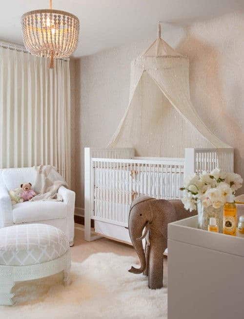 A boho chandelier hangs over the round ottoman sitting on a white faux fur rug over carpet flooring. There are white armchair and skirted crib with a canopy net overhead.