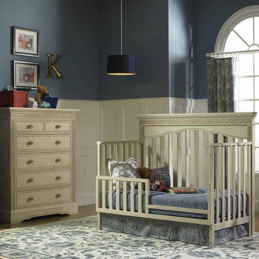 A drum pendant light illuminates this classic nursery showcasing a skirted crib and drawer chest accented with black framed wall arts. It has a floral area rug and patterned drapes covering the arched window.