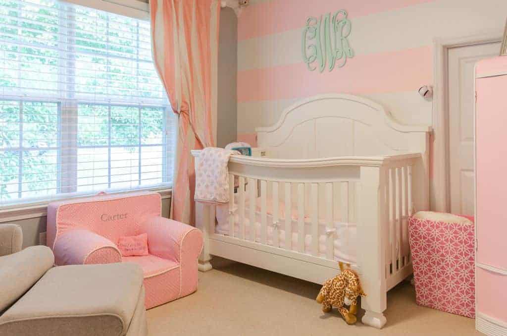 Airy nursery features a pink dotted mini armchair and a white crib placed against the striped accent wall. It has carpet flooring and glazed windows bringing natural light in.