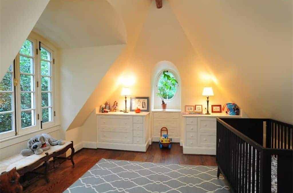 Warm nursery with vaulted walls and hardwood flooring topped by a gray patterned rug. It includes a dark wood crib and white cabinets lighted by traditional table lamps.